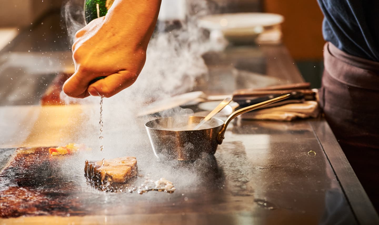 鉄板の上で豪快に焼かれる魚料理と立ち上る熱い湯気、弾ける脂の躍動感あふれるシズルカット