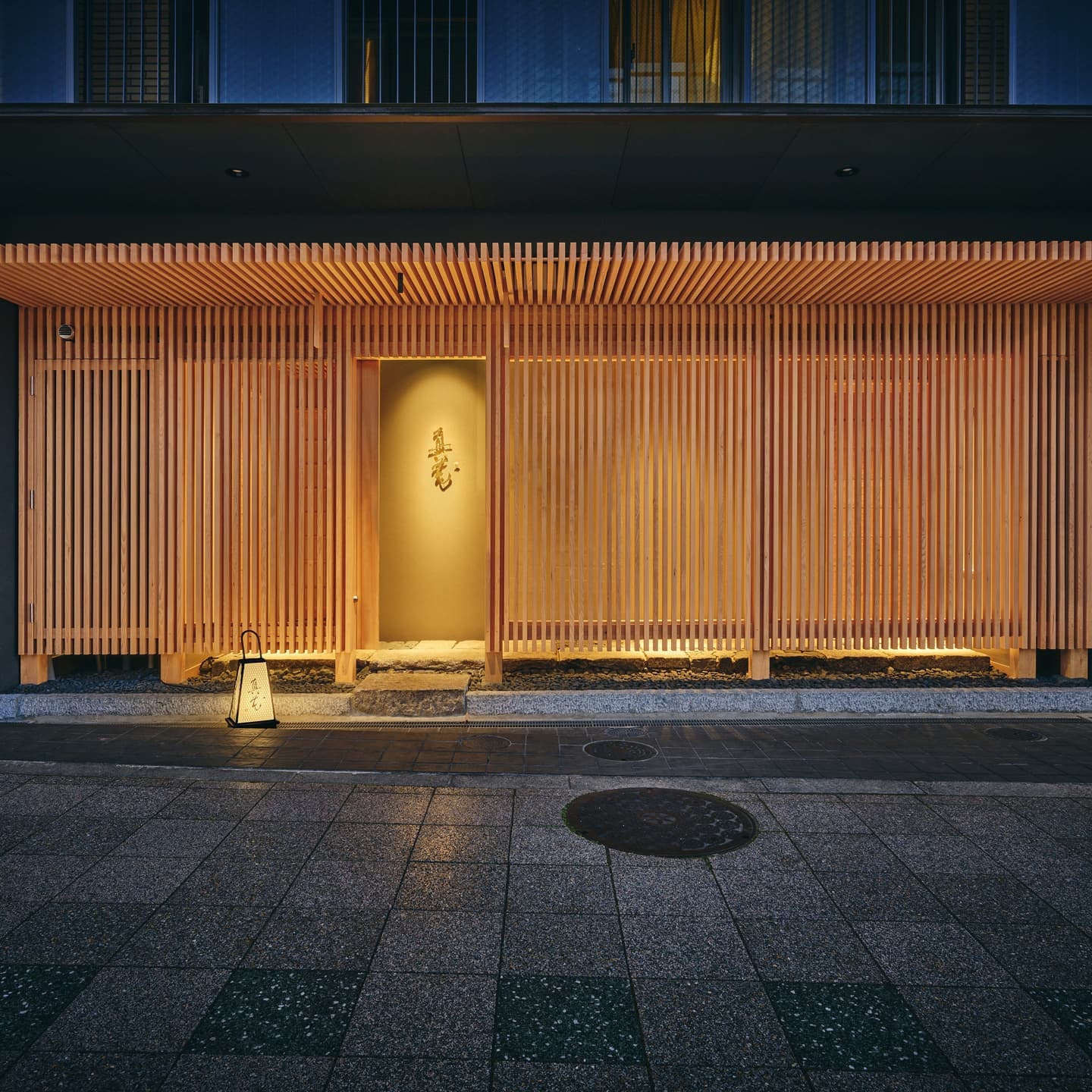 Alt設定値： 夕暮れ時に温かな光が漏れる和モダンの木製格子に包まれた店舗外観、計算されたシンメトリーと陰影が美しい建築撮影