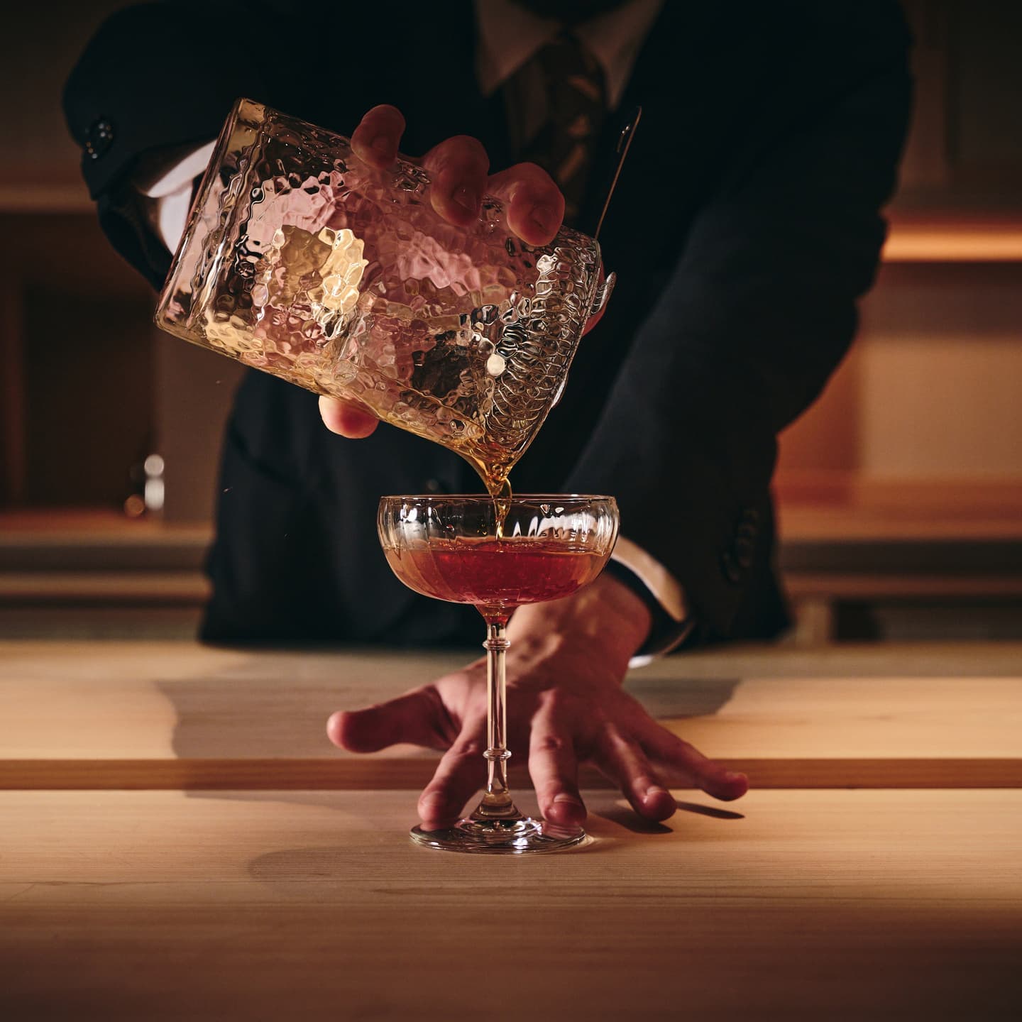 ミキシンググラスからカクテルを注ぐバーテンダーの鮮やかな手元、琥珀色の液体と氷の輝きが交差するドラマチックなバーシーン撮影