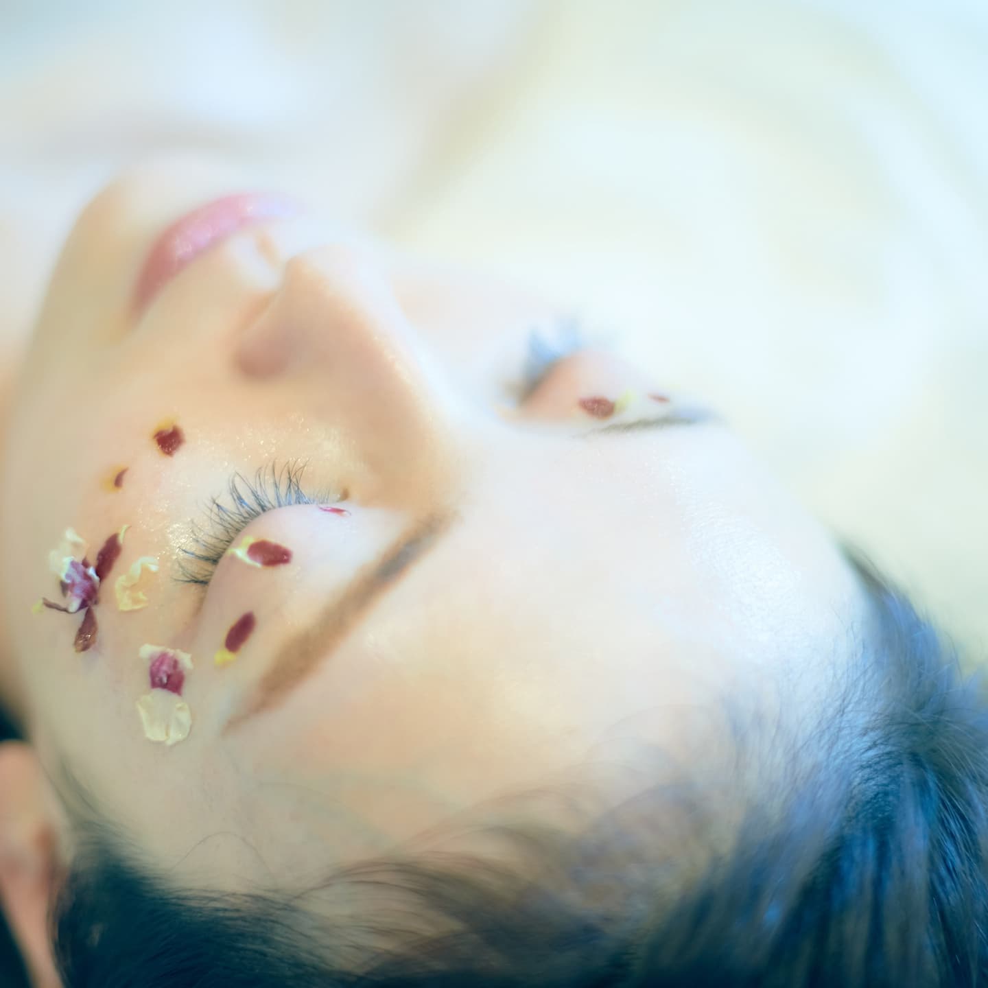 目元に花びらをあしらい、安らかな表情で横たわる女性のクローズアップ。肌の質感と生命力を強調し、美容やライフスタイルの物語を想起させるビジュアル。