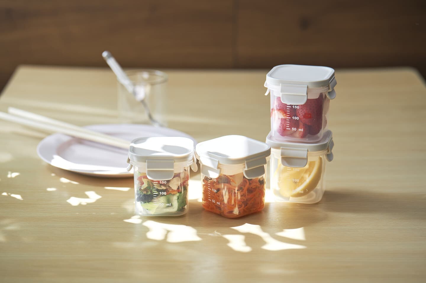 大阪のスタジオで撮影された、柔らかな木漏れ日が差し込む食卓の風景。彩り豊かな食材を入れた透明な保存容器を、ライフスタイルの一部として情緒的に表現。