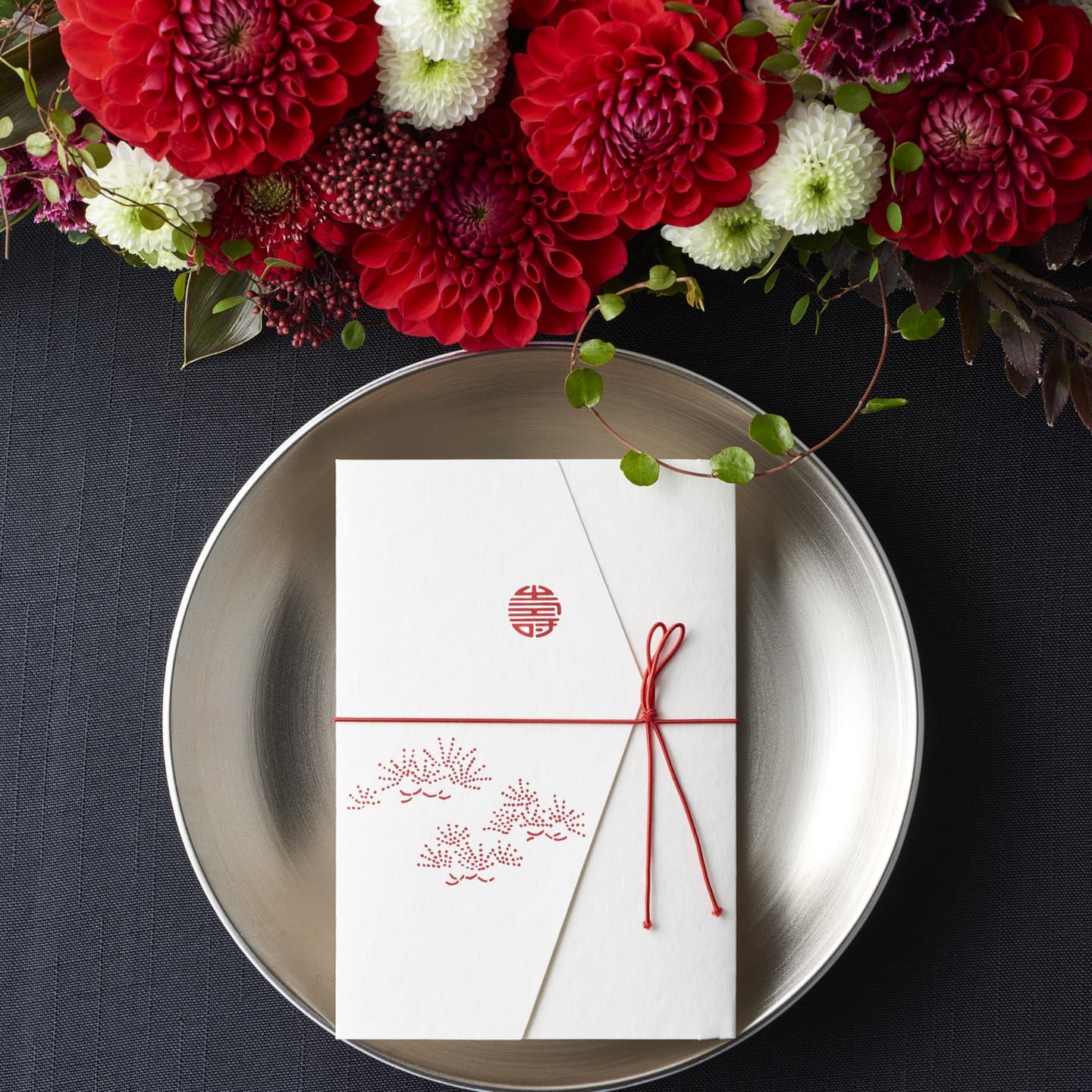 大阪のスタジオで撮影された、鮮やかな赤いダリアと洗練されたデザインののし袋。俯瞰（真上）からのアングルで、紙の柔らかな質感と銀色の皿、花の瑞々しさを対比させた、ギフト・ステーショナリーブランド向けの静物写真。