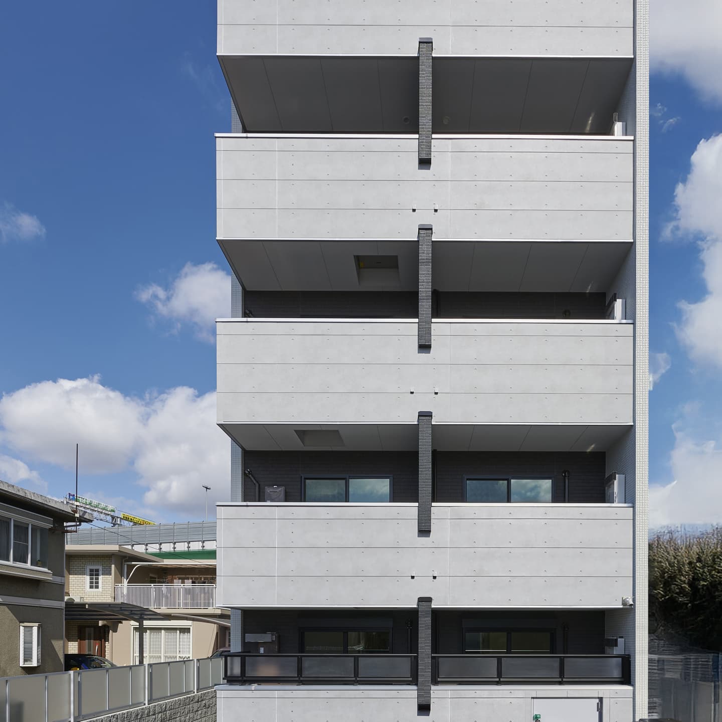 コンパクトレジデンス（賃貸マンション）の外観竣工写真。青空を背景に、コンクリートの質感と垂直水平のラインを強調したシャープな建築ポートフォリオ。