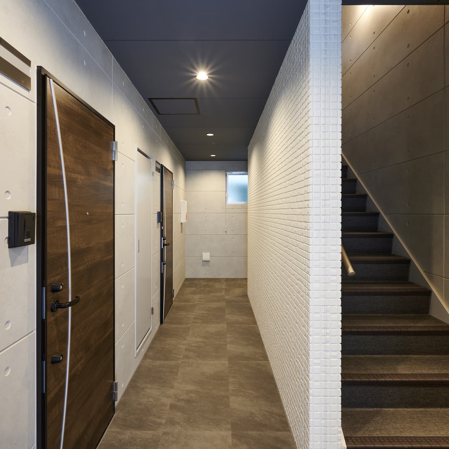 マンション共用部の内覧写真。コンクリート打ち放しの壁面と木目調のドアが調和する廊下を、奥行きを感じさせる構図で捉えた竣工ビジュアル。