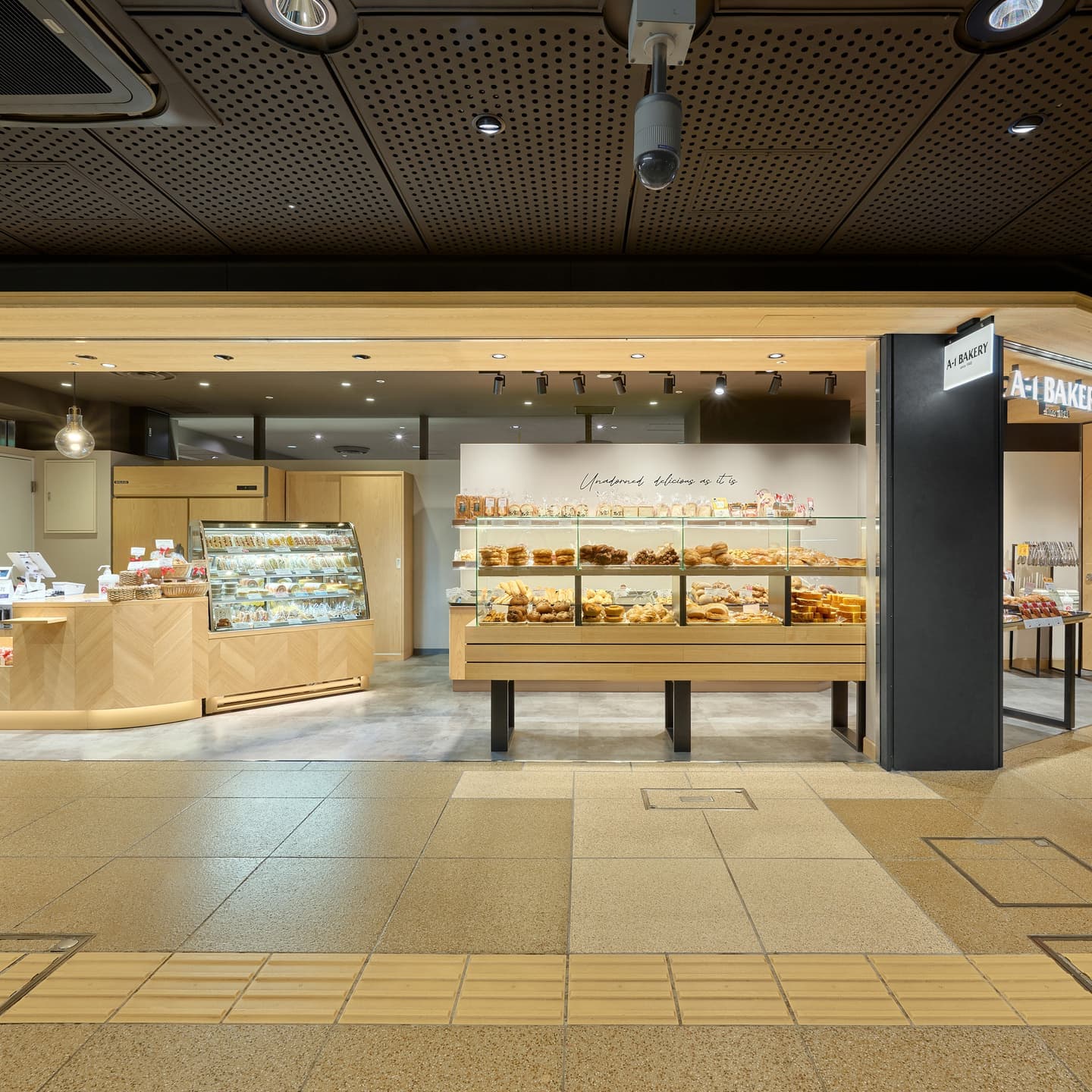 開放的な間口にヘリンボーン柄のカウンターを配したA-1 BAKERYの店舗。ガラスケース越しに並ぶパンと、温白色のスポットライトが演出する賑やかな雰囲気。